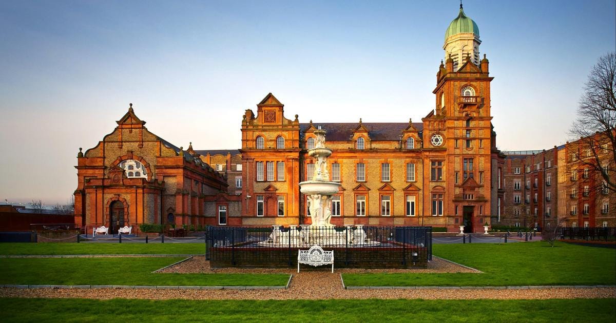 A historic brick building with an ornate clock tower and dome, featuring a large central fountain in the foreground surrounded by a well-manicured lawn and pathway