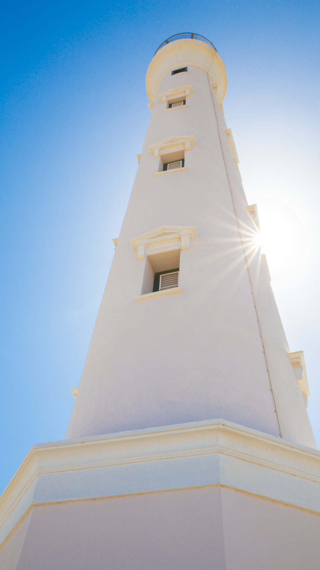 Up-close looking at a tall very white California lighthouse in Aruba with clear blue skies above.