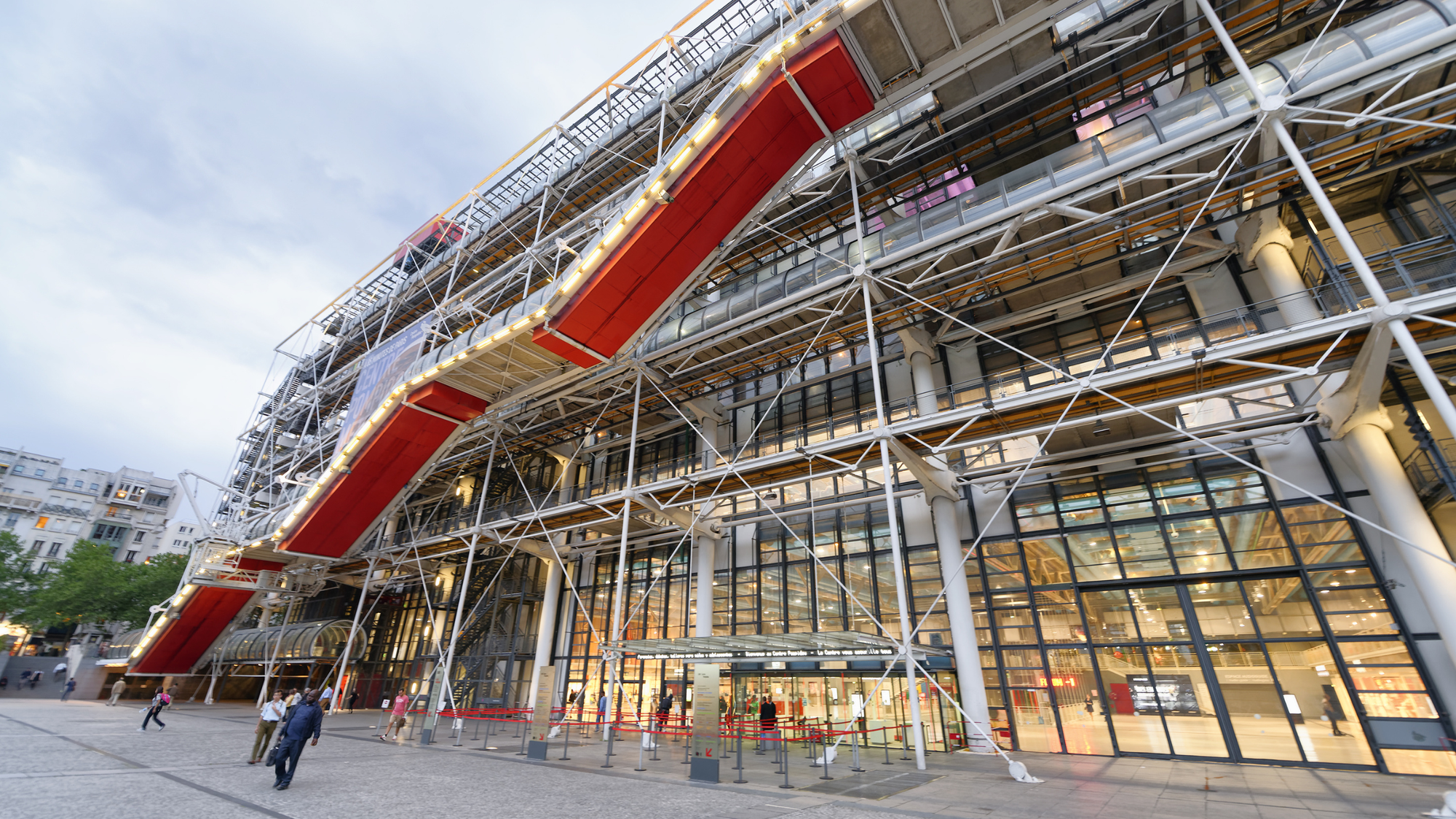 A large, futuristic building with an industrial design, the Centre Pompidou features an exterior of exposed steel beams, colorful pipes, and ducts in bright shades of blue, red, green, and yellow. The front of the building has a prominent glass-enclosed escalator that zigzags up the facade, leading to the upper floors. 