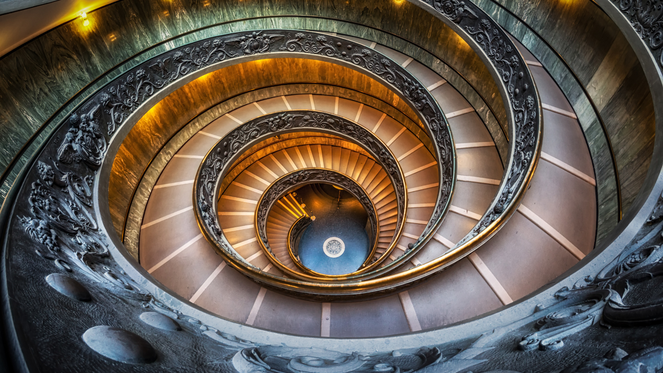 A spiraling, circular staircase inside the Vatican Museum, featuring ornate balustrades and intricate design elements. The staircase has a grand, sweeping appearance, with each level descending in a symmetrical, helix-like fashion. Light streams in from above, highlighting the elegant structure and its detailed craftsmanship.