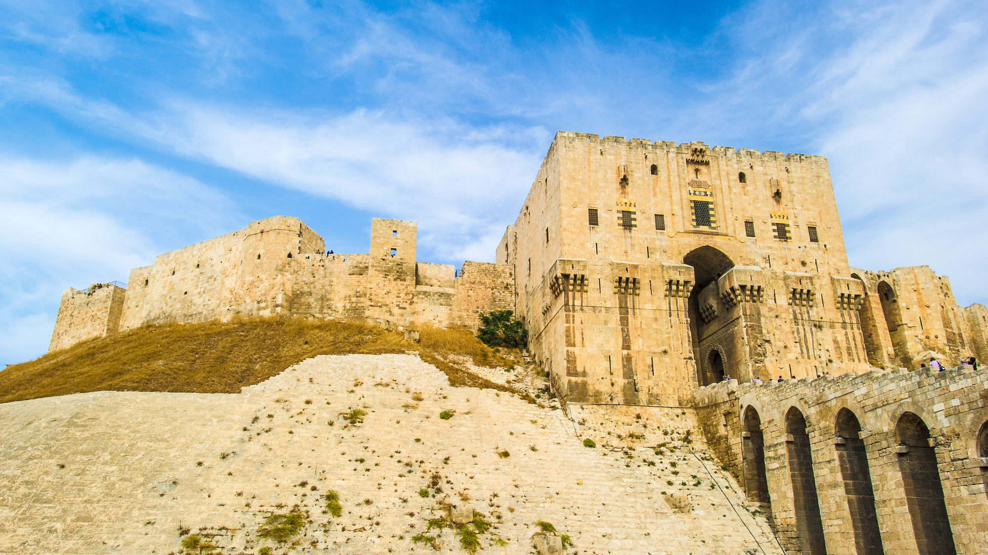 Citadel of Aleppo in Syria, a massive ancient fortress with towering stone walls perched on a hill.