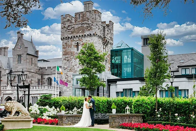 A bride and groom standing in front of Clontarf Castle in Dublin, surrounded by a lush garden with vibrant flowers and greenery. The historic stone architecture of the castle towers behind them, complemented by a modern glass extension, under a bright blue sky with fluffy white clouds.