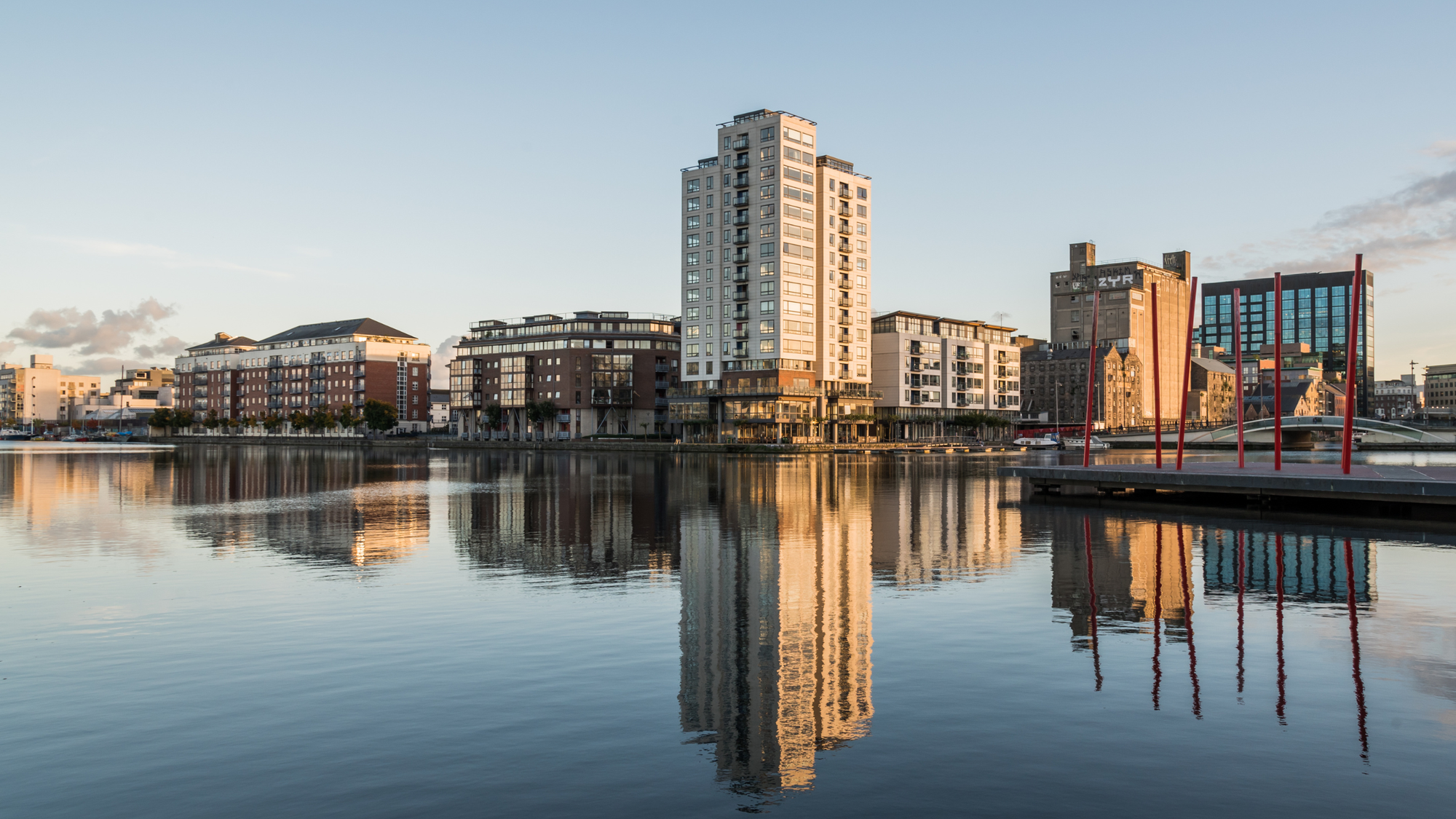 View of the Docklands neighborhood in Dublin, featuring modern high-rise buildings and sleek architecture reflected in the calm water of the River Liffey, with a clear sky and red poles adding to the urban landscape.