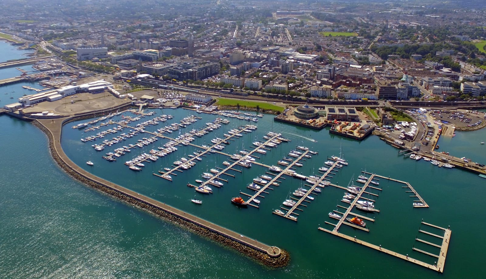 Aerial view of Dun Laoghaire neighborhood in Dublin, showcasing a bustling marina filled with boats and yachts, surrounded by the town's buildings and coastline. The image highlights the expansive harbor and the vibrant waters of Dublin Bay.