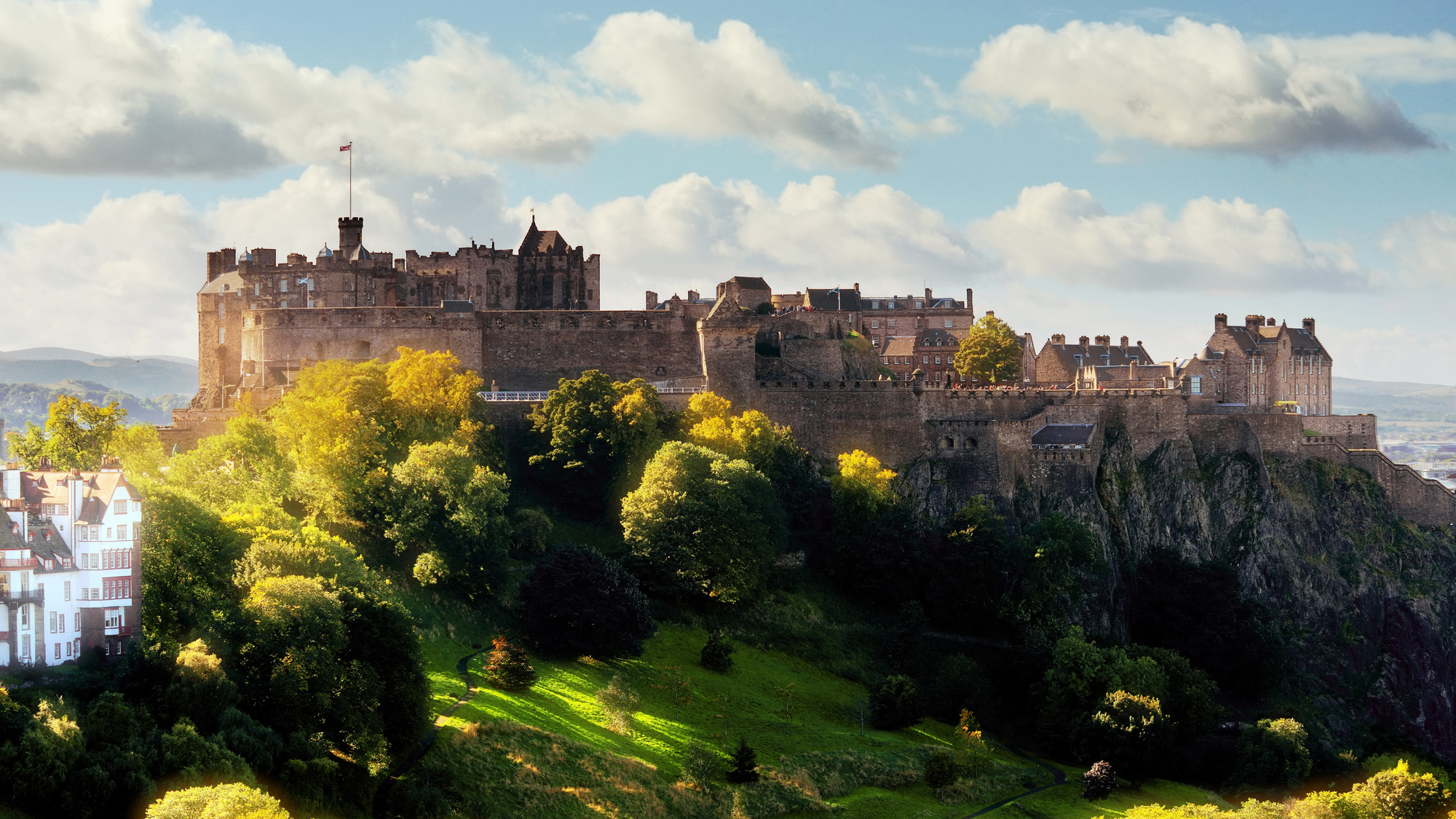 Edinburgh Castle in Scotland, perched on a green hill with scattered trees, under partly cloudy skies.