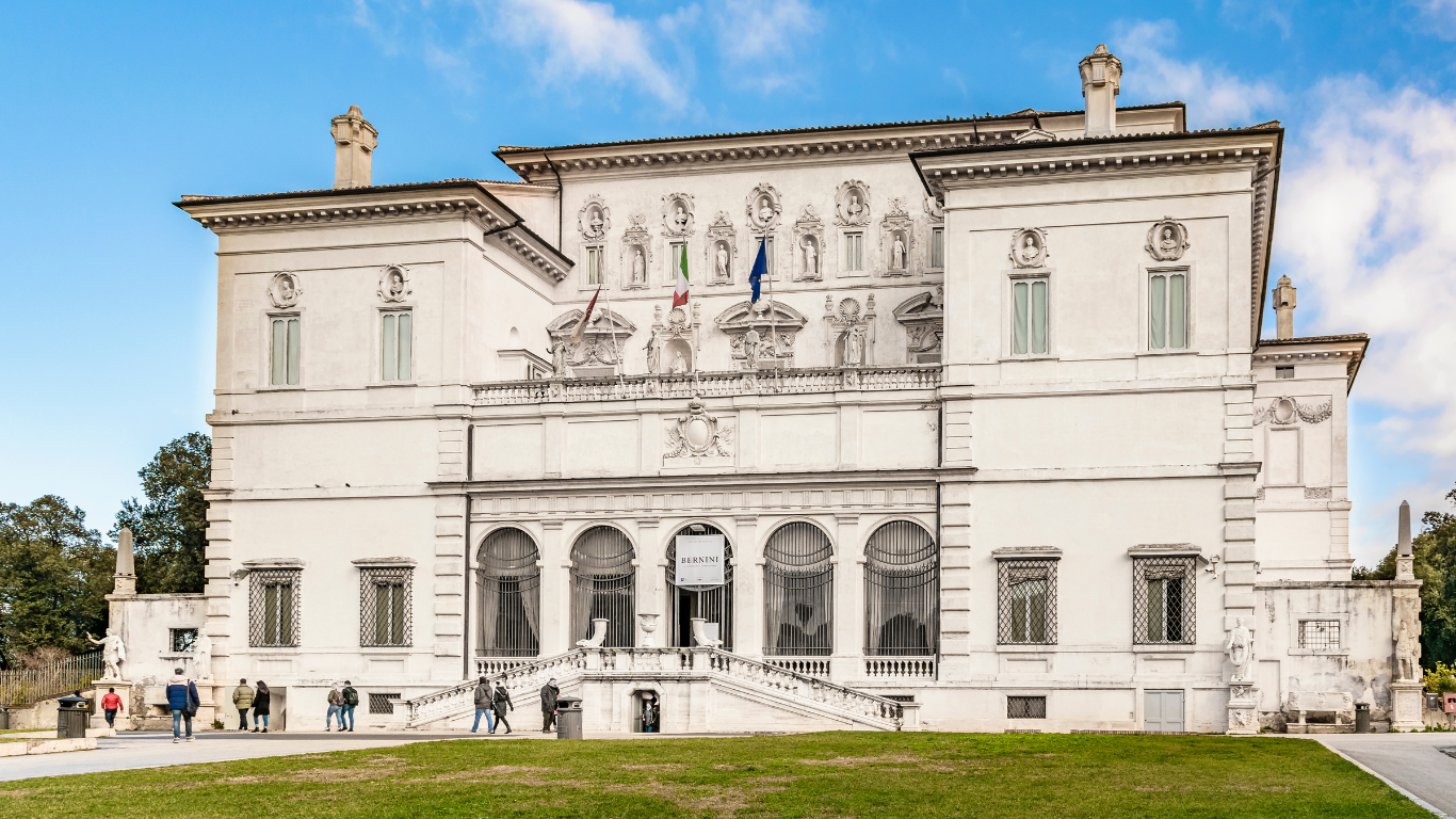 a grand villa set within the lush Borghese Gardens in Rome. The building features classical Italian architecture with a symmetrical facade, adorned with sculptures, decorative reliefs, and arched windows. The villa is surrounded by manicured gardens, adding to its elegance. The entrance is framed by a set of steps leading up to the main door, reflecting the rich history and artistic significance of the gallery within.