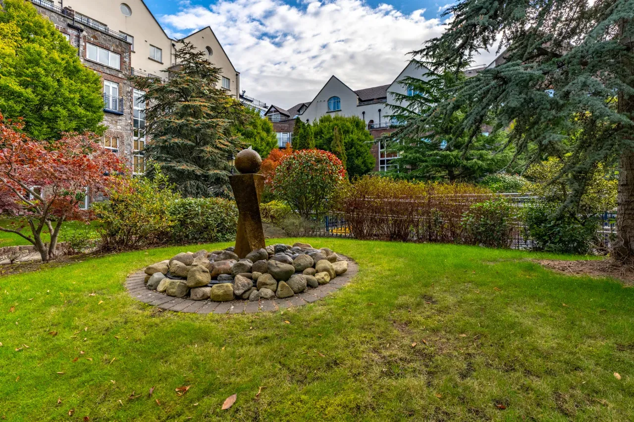 Grace Manor Park in the Drumcondra neighborhood of Dublin, featuring a modern stone sculpture surrounded by a circular arrangement of rocks and lush greenery. The background showcases residential buildings with a mix of colorful trees and plants under a partly cloudy sky