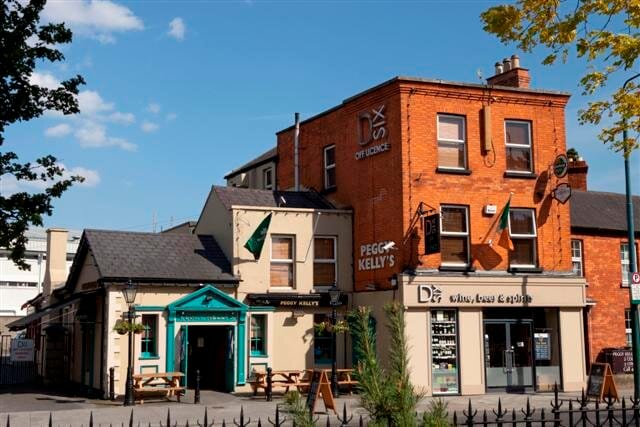 Exterior view of Pegg Kelly's pub in Harold's Cross, Dublin, Ireland. The pub features traditional Irish architecture with a vibrant green entrance, and an adjoining building houses an off-license store. The scene is set on a sunny day with a clear blue sky.