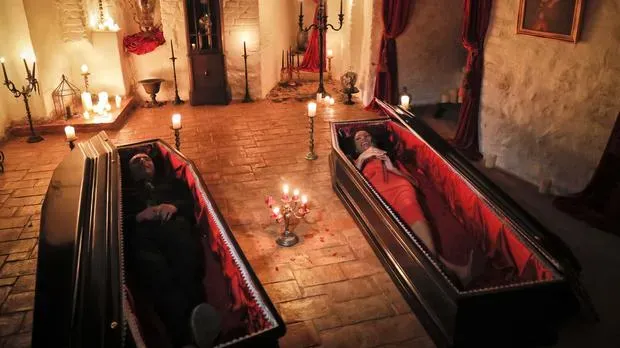 a male Dracula and female vampire mannikin laying in coffins with the lids off and dressed in formal attire and surrounded by lit candles