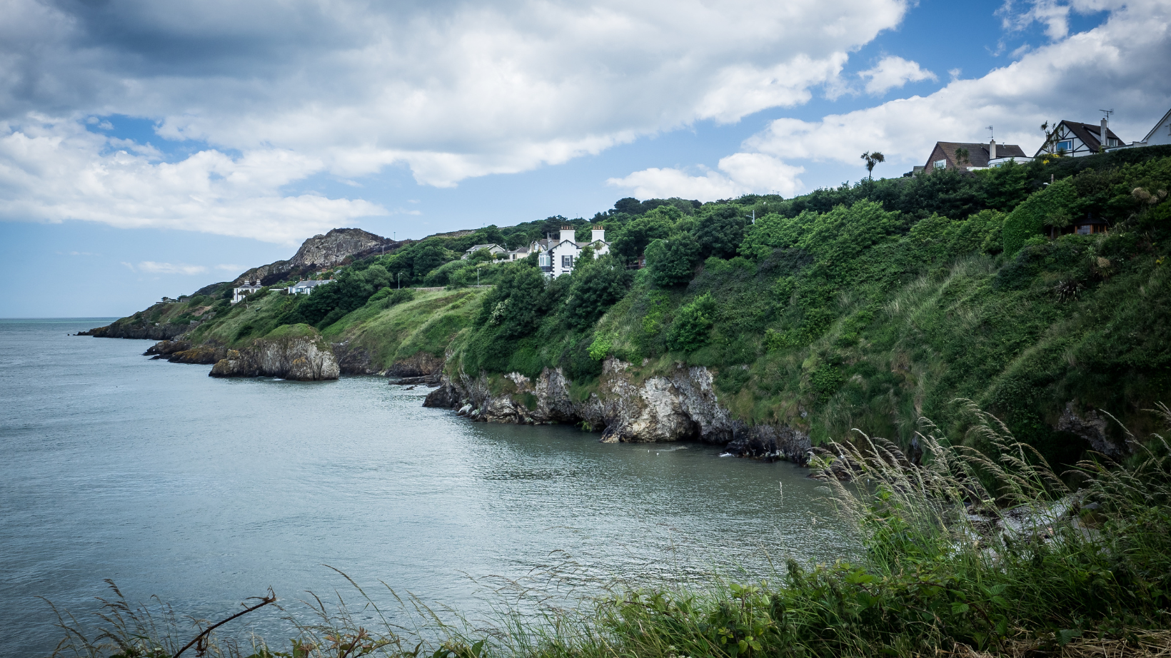 Scenic coastal view of Howth Village near Dublin, featuring lush green cliffs overlooking the calm sea. A few houses are perched on the hillside, offering a serene and picturesque landscape under a partly cloudy sky.