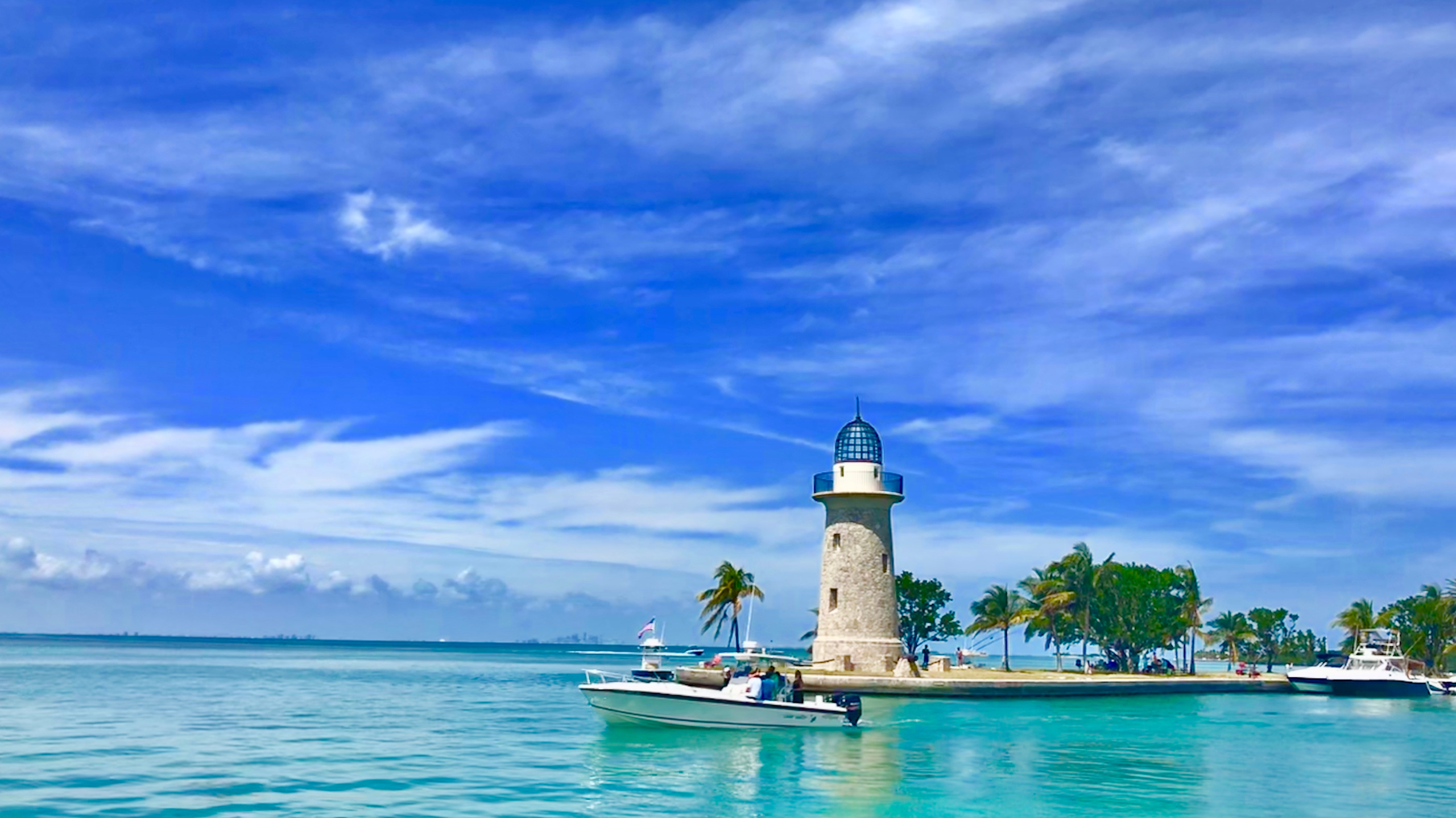 Scenic view of the Cape Florida Lighthouse on Key Biscayne, Miami, with crystal clear waters, boats, and palm trees under a vibrant blue sky.