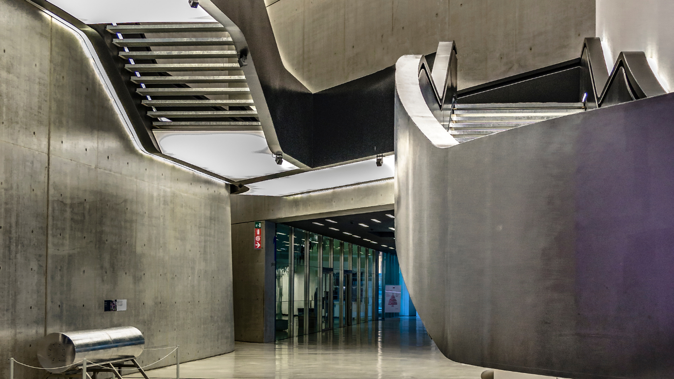 The interior of MAXXI – National Museum of 21st Century Art, featuring sleek, modern staircases with clean lines and minimalist design. The black, floating staircases twist and turn in a dynamic, architectural style against the white walls of the building. The open, airy space is filled with natural light streaming from large windows, emphasizing the contemporary and avant-garde atmosphere of the museum's design.