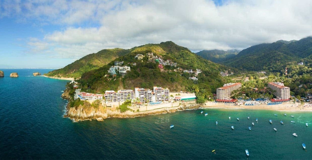 seaside resorts with boats in the water next to white sandy shores that are surrounded in mountains covered in thick jungle and trees in Mismaloya Beach in Puerto Vallarta 