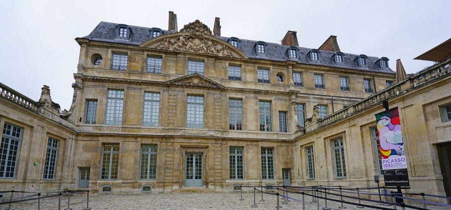 The building features a light stone facade with ornate architectural details, including large windows, decorative carvings, and wrought-iron balconies. 