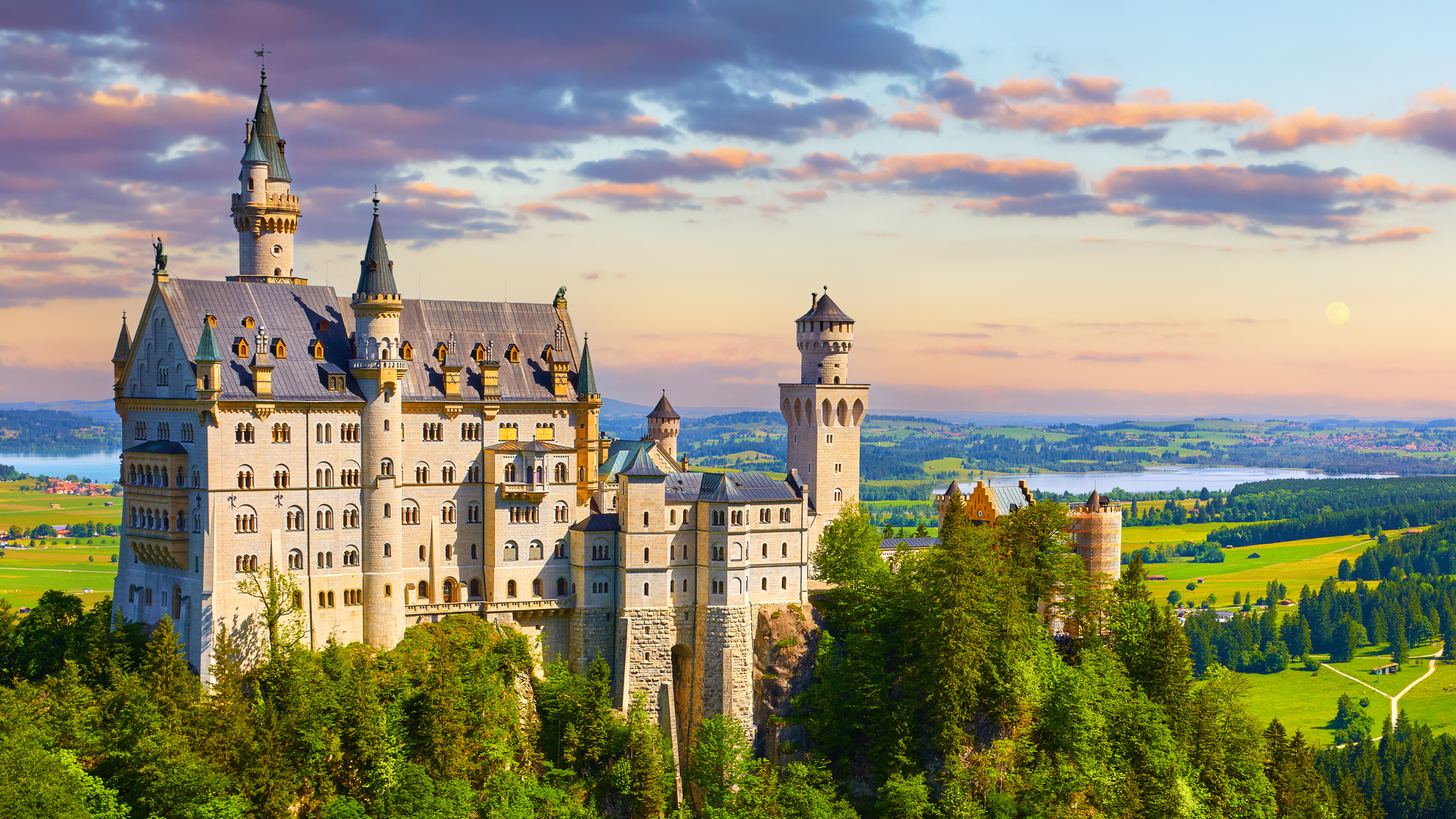 Neuschwanstein Castle in Germany, with its fairytale-like towers and white stone walls, surrounded by green pine trees and grassy fields on a sunny summer day.