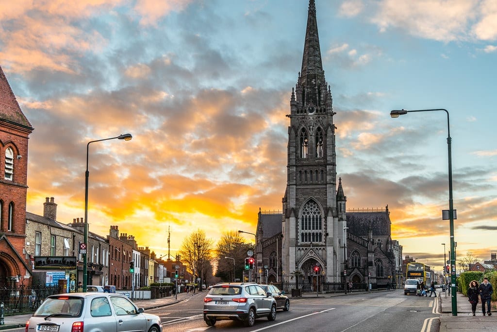 Phibsborough neighborhood in Dublin, featuring a striking church with a tall spire at sunset. Cars and pedestrians are seen on the street, with a vibrant sky of orange and blue hues in the background