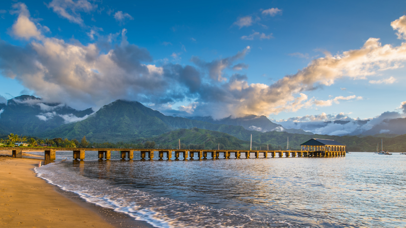 Pier going out into the ocean with mountains in the background with partly cloudy skies in Hanalei, Hawaii.