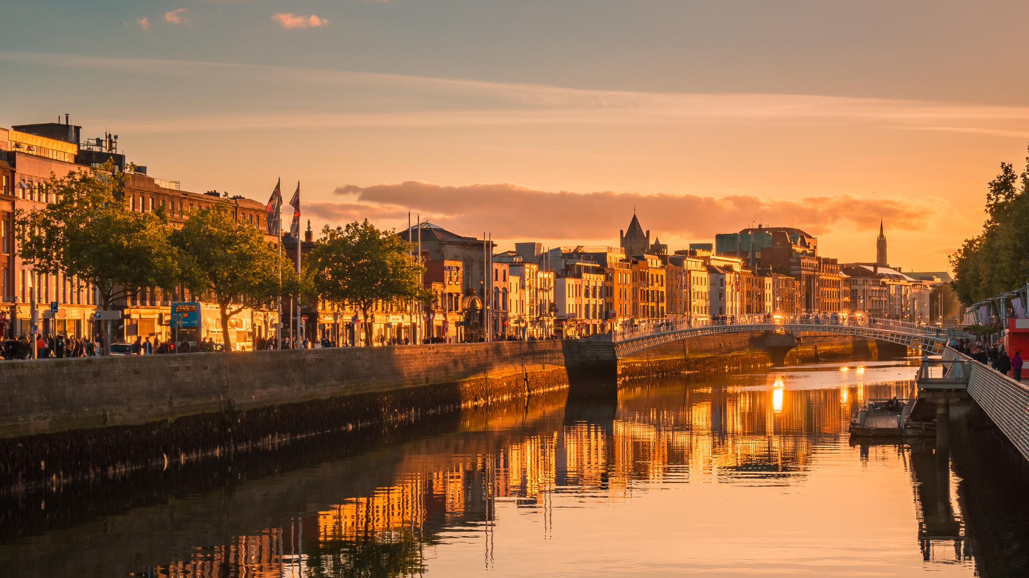 View of the Portobello neighborhood in Dublin at sunset, with the Ha'penny Bridge crossing the River Liffey. The golden light illuminates the historic buildings and the waterfront, creating a warm and picturesque atmosphere.