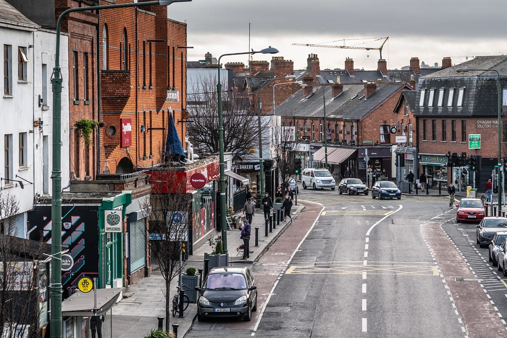 A bustling street in the Ranelagh Village neighborhood of Dublin, Ireland, lined with red-brick buildings housing shops, cafes, and businesses. People are walking along the sidewalk, and cars are parked and driving on the road, reflecting the vibrant, urban atmosphere of the area.