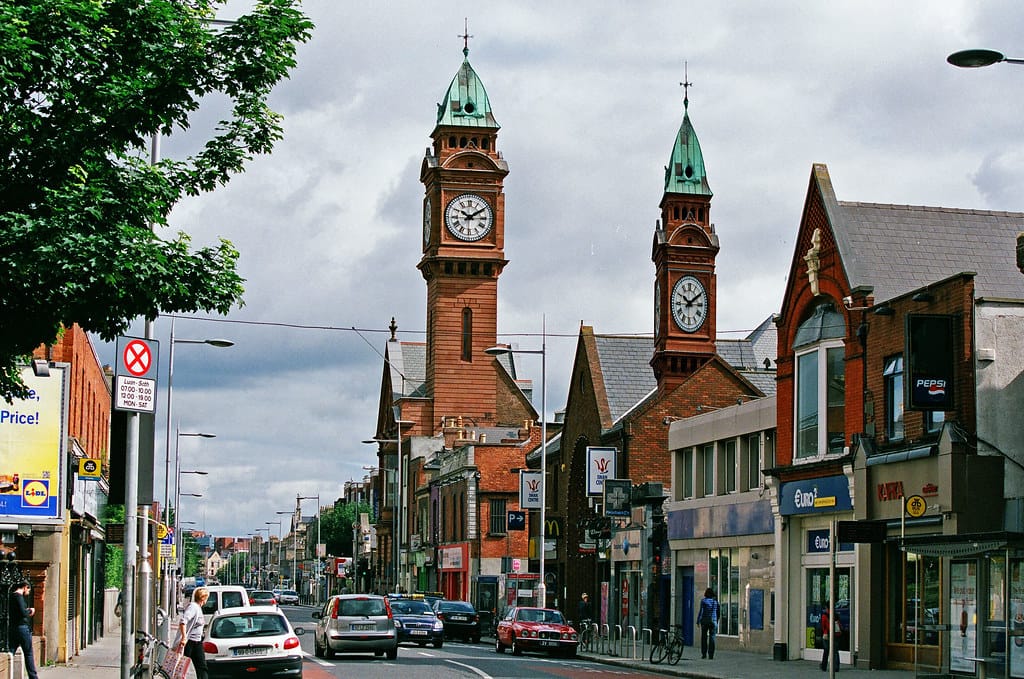 Street view of the Rathmines neighborhood in Dublin, Ireland, featuring the iconic red-brick clock towers with green domes. The street is bustling with cars and pedestrians, lined with shops and businesses under a cloudy sky.