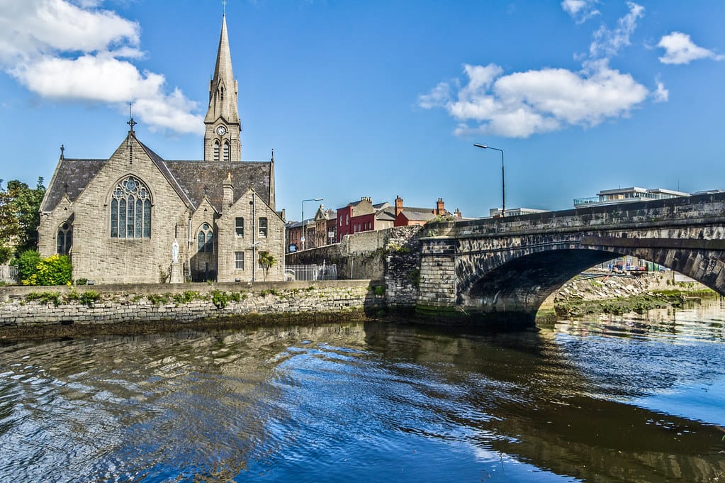 View of a St. Patrick's historic church and stone bridge in the Ringsend neighborhood of Dublin, Ireland. The church's pointed spire and Gothic architecture are reflected in the calm water of the river, with a clear blue sky and scattered clouds overhead, creating a serene urban scene.