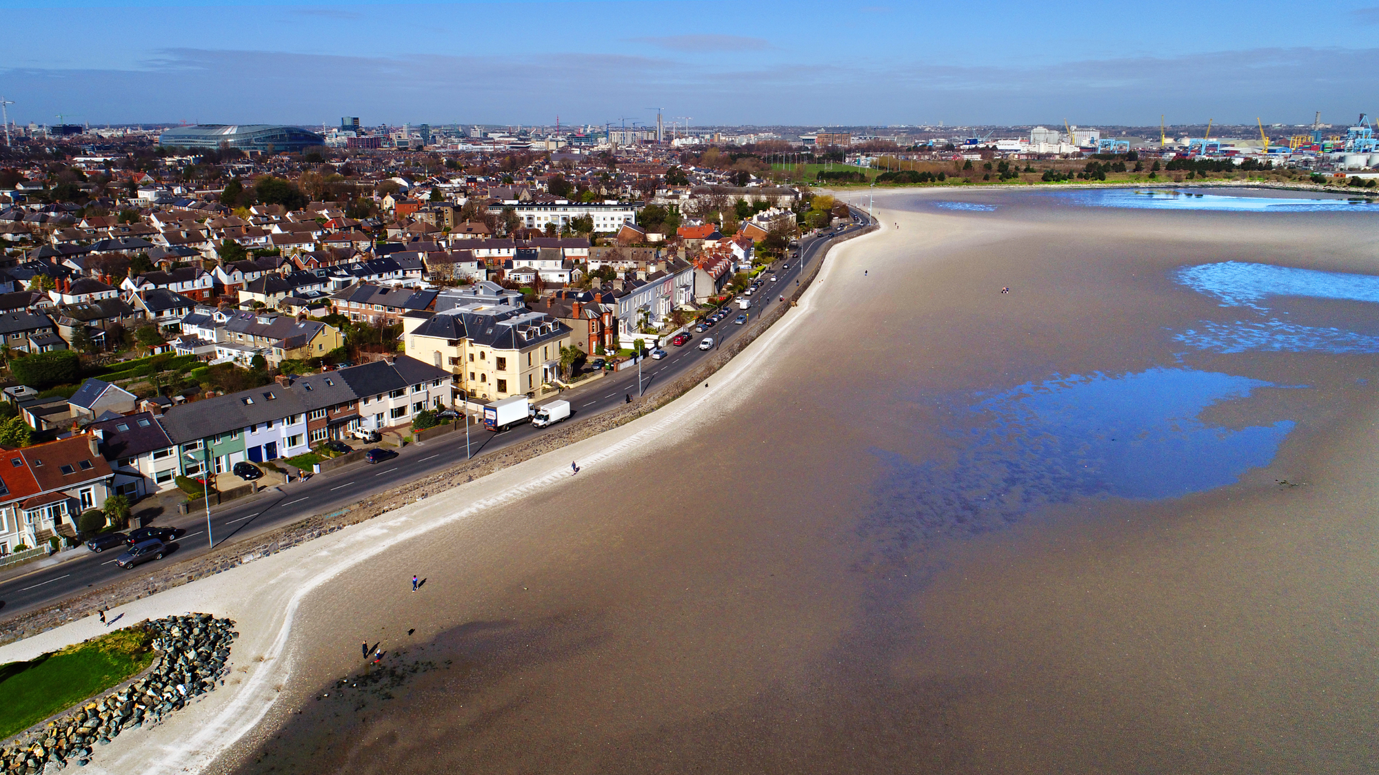 Aerial view of the Sandymount neighborhood in Dublin, featuring a stretch of sandy beach and a scenic promenade along the coastline. The neighborhood showcases rows of charming houses with the cityscape and a stadium visible in the background under a clear blue sky.