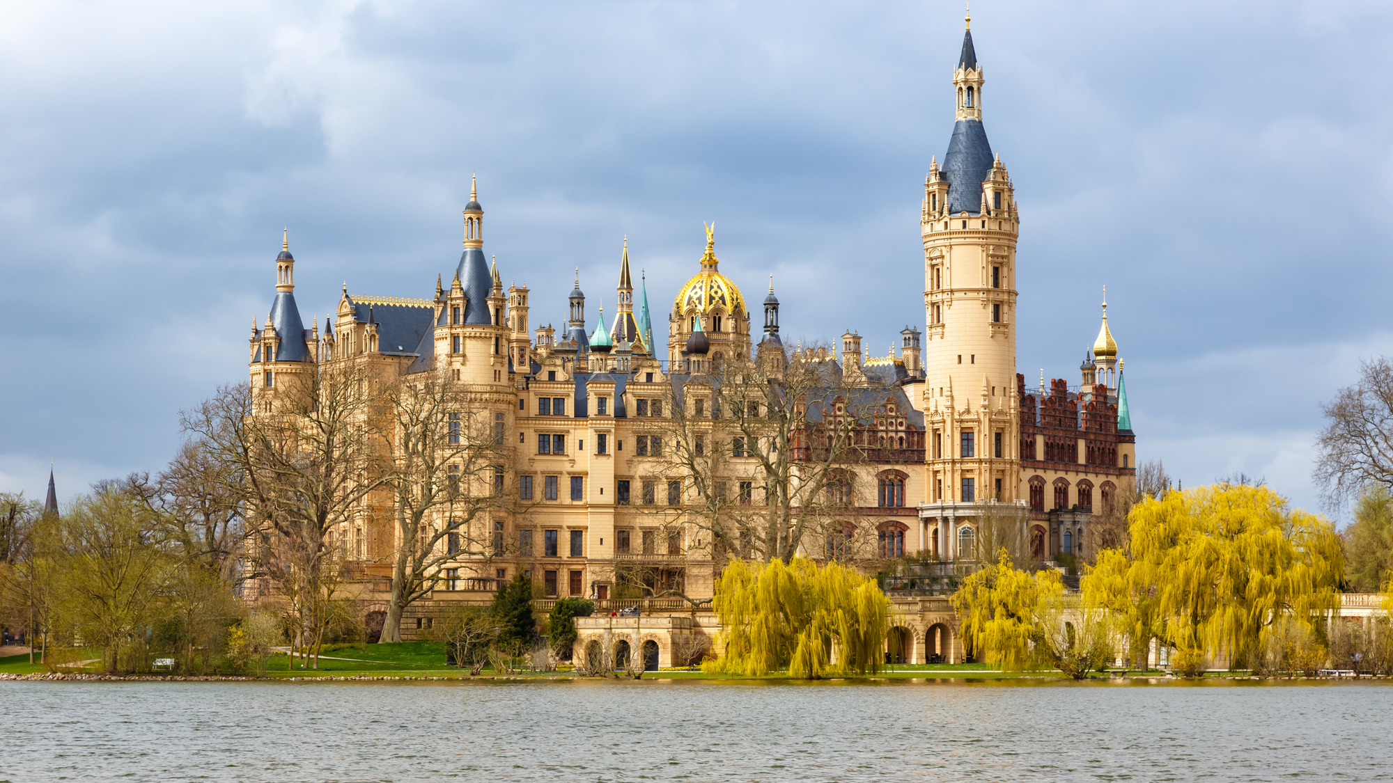 Schwerin Castle in Germany, with its golden spires and ornate towers, reflected in the lake, surrounded by early fall trees with hints of autumn color.