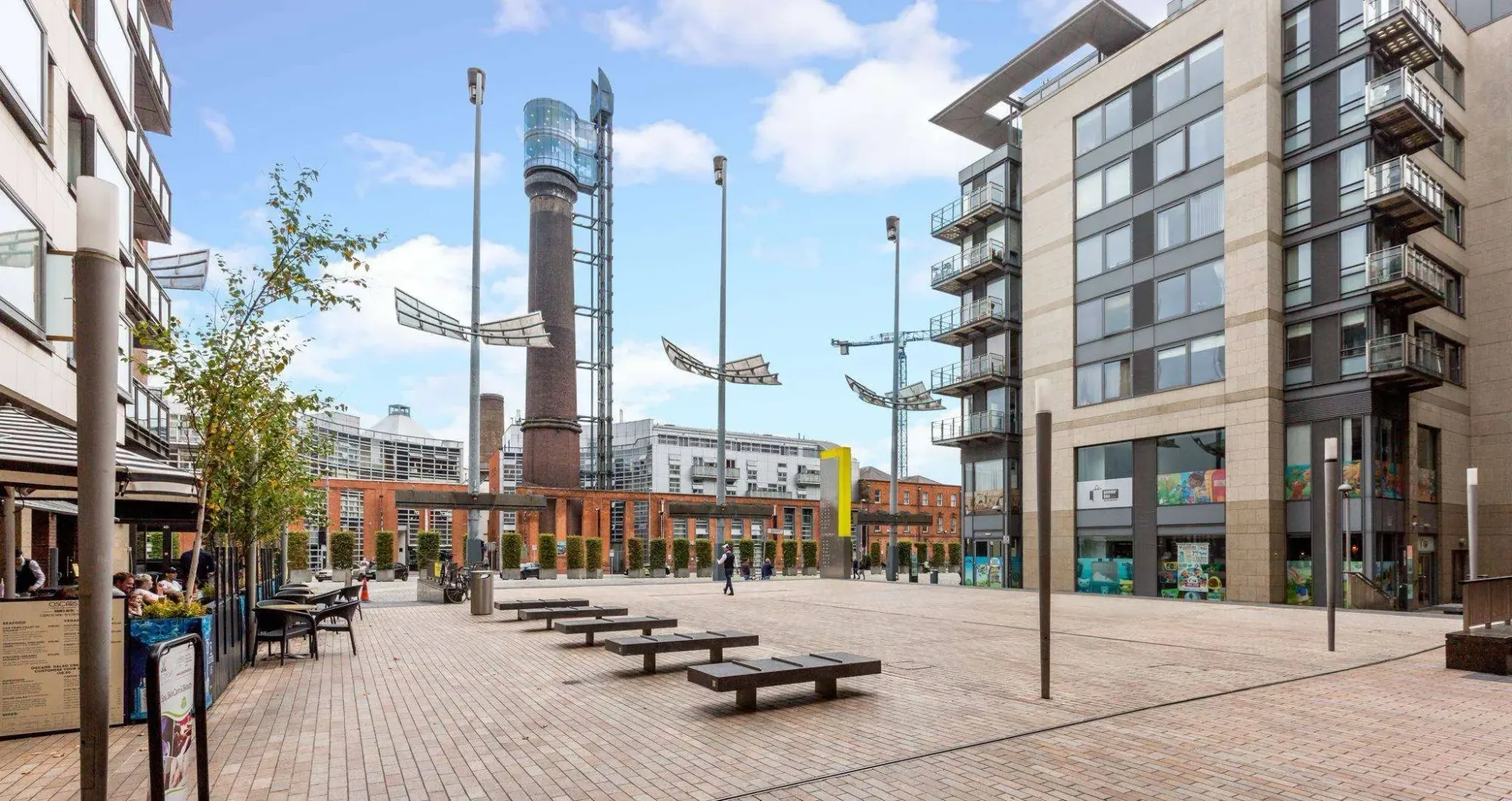 A modern urban square with brick pavement, surrounded by contemporary buildings. A tall, cylindrical tower and unique, sail-like street lights are prominent in the center. Outdoor seating and benches are scattered throughout the area