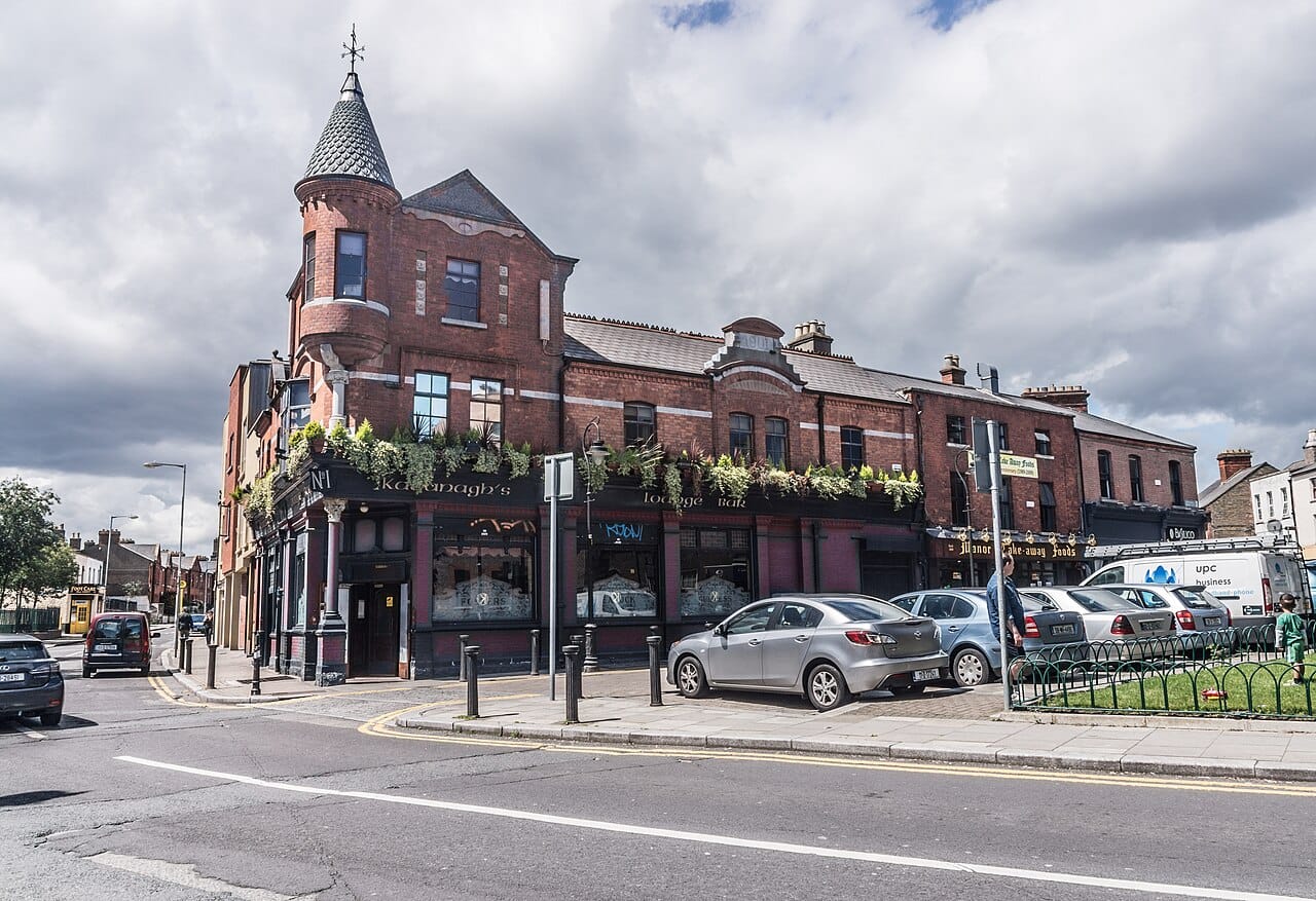 Stoneybatter neighborhood in Dublin, showcasing a historic red-brick building with a traditional pub on the corner, surrounded by parked cars and a small green space, under a partly cloudy sky