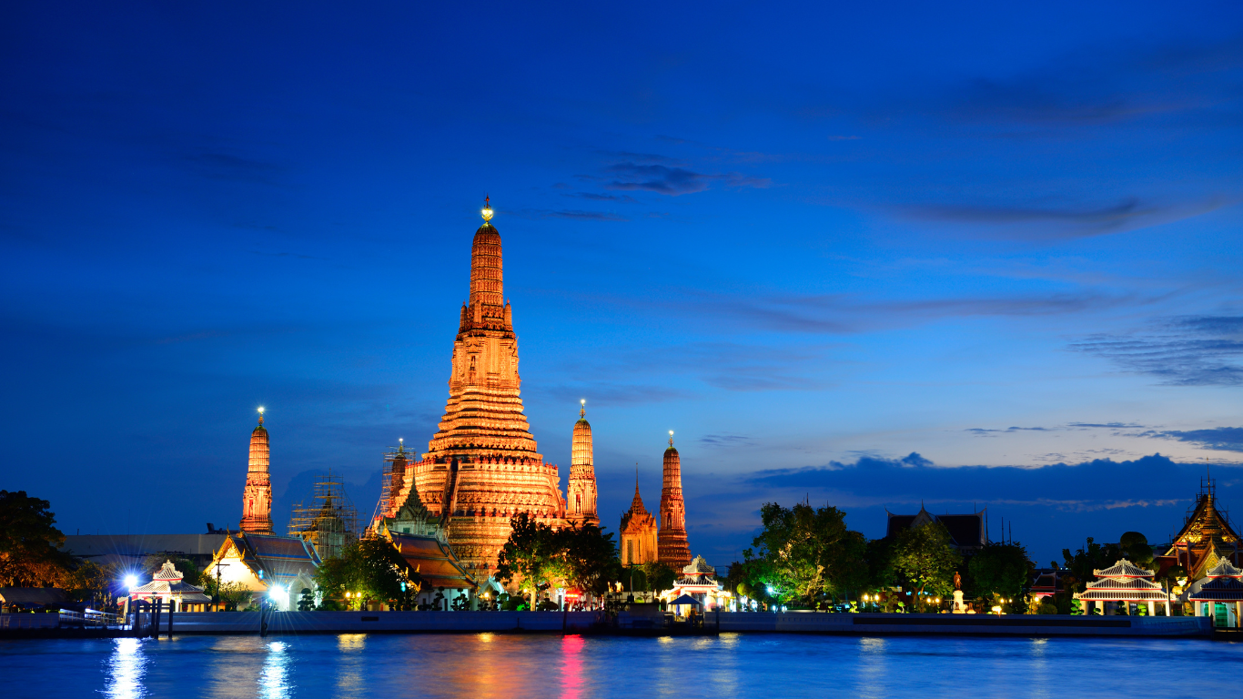 Nighttime with lights lighting up a beautiful temple that has 4 surrounding structures next to a river running through the city of Bangkok