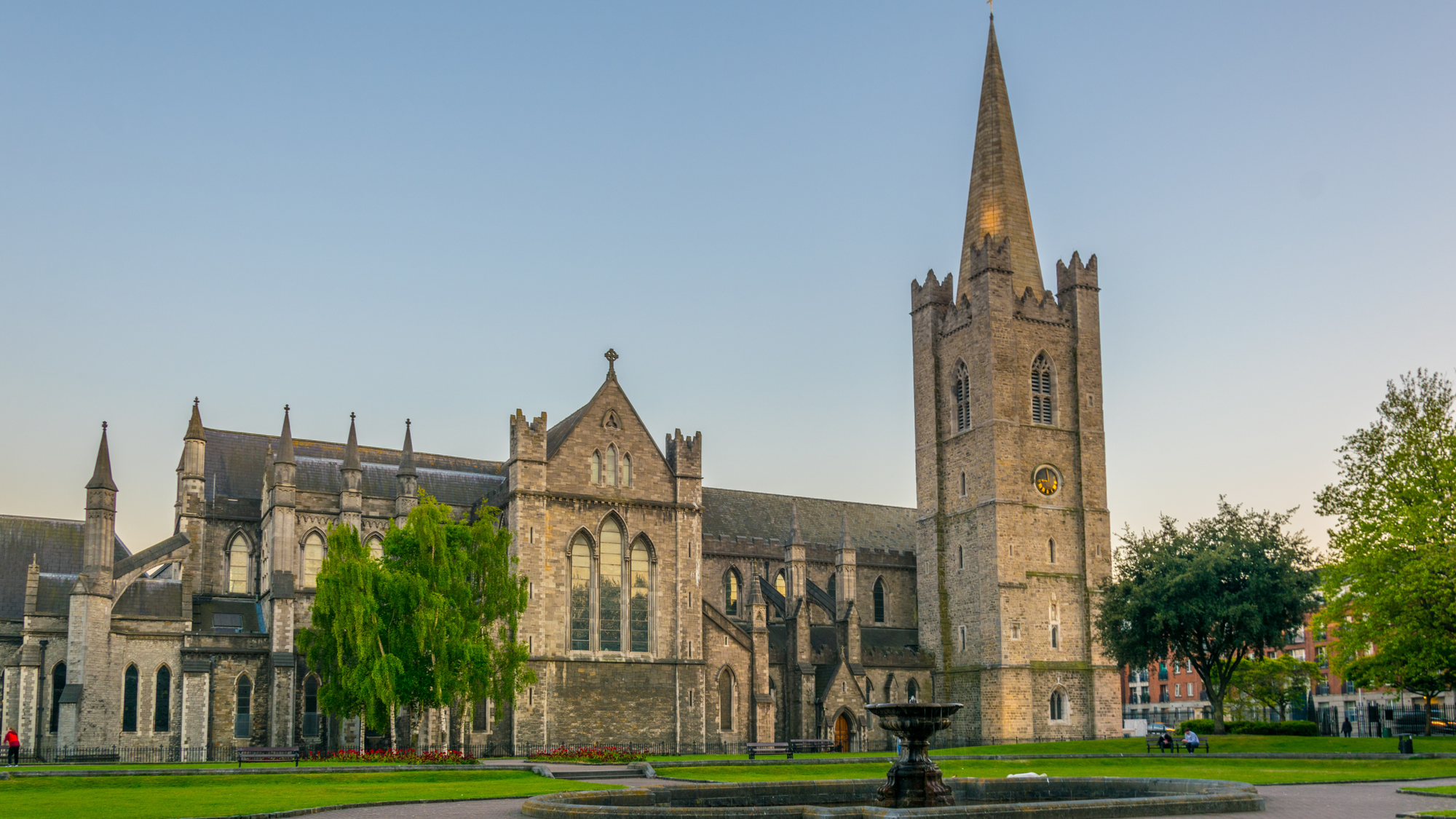 St. Patrick's Cathedral in The Liberties, Dublin, featuring the historic stone architecture and tall spire of the cathedral under a clear sky. The foreground shows a lush green park area with a fountain and people sitting on benches, adding to the serene atmosphere.