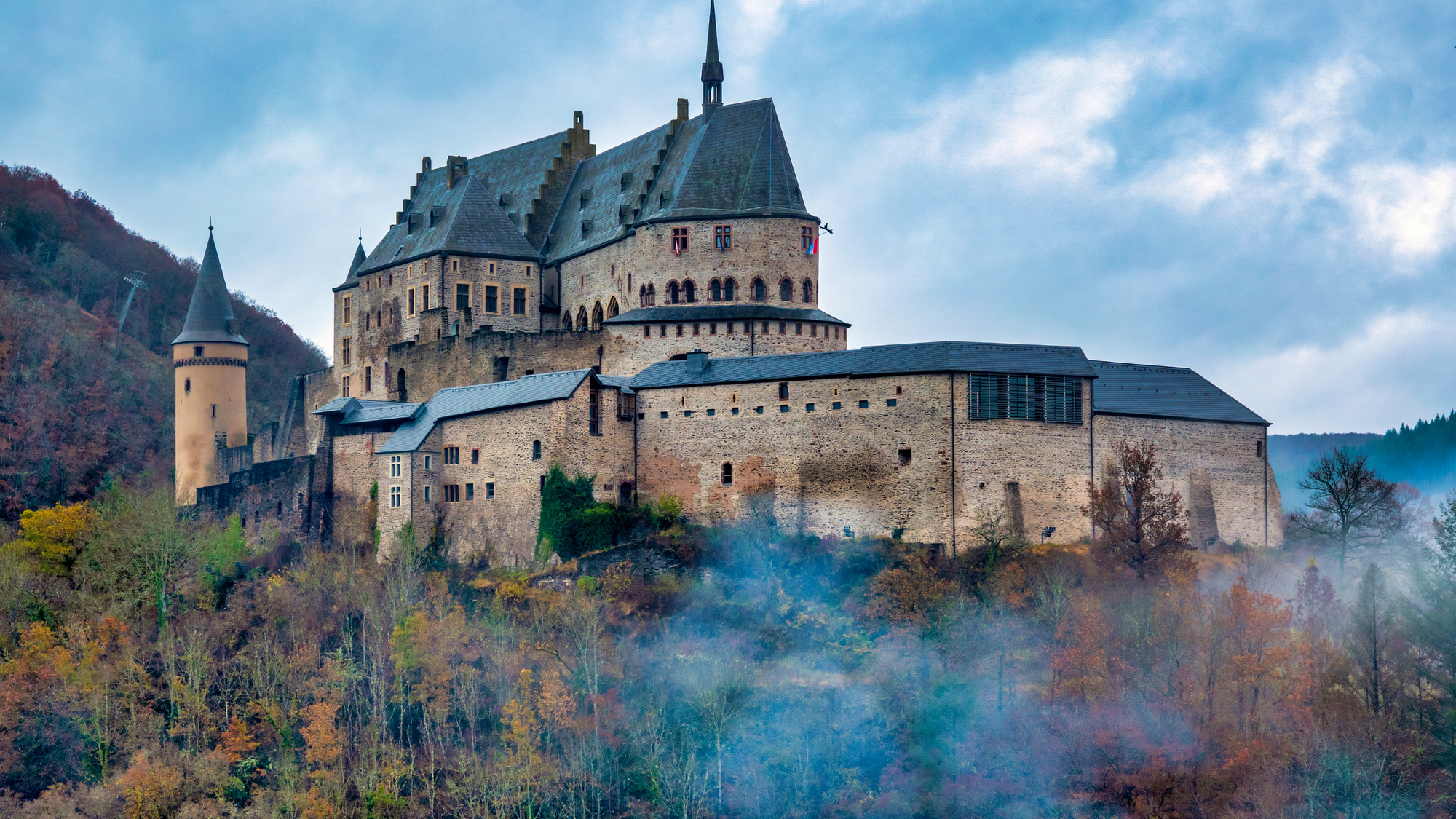 Vianden Castle in Luxembourg, with its tall stone towers emerging through the fog, surrounded by autumn-colored trees