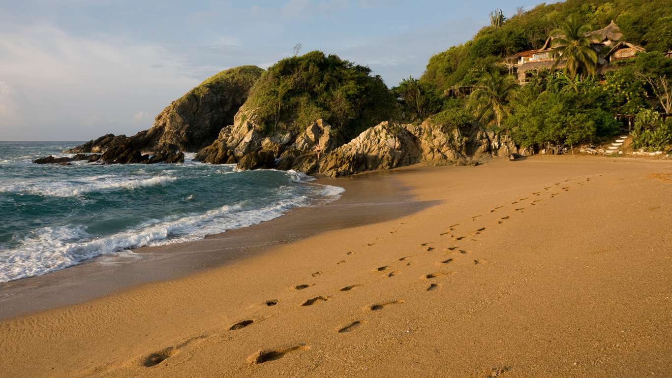 waves crashing the shores of the sand with large rocks with foliage making up a beach enclave, and feet prints in the sand going to a stair case leading up the mountainside to a resort 