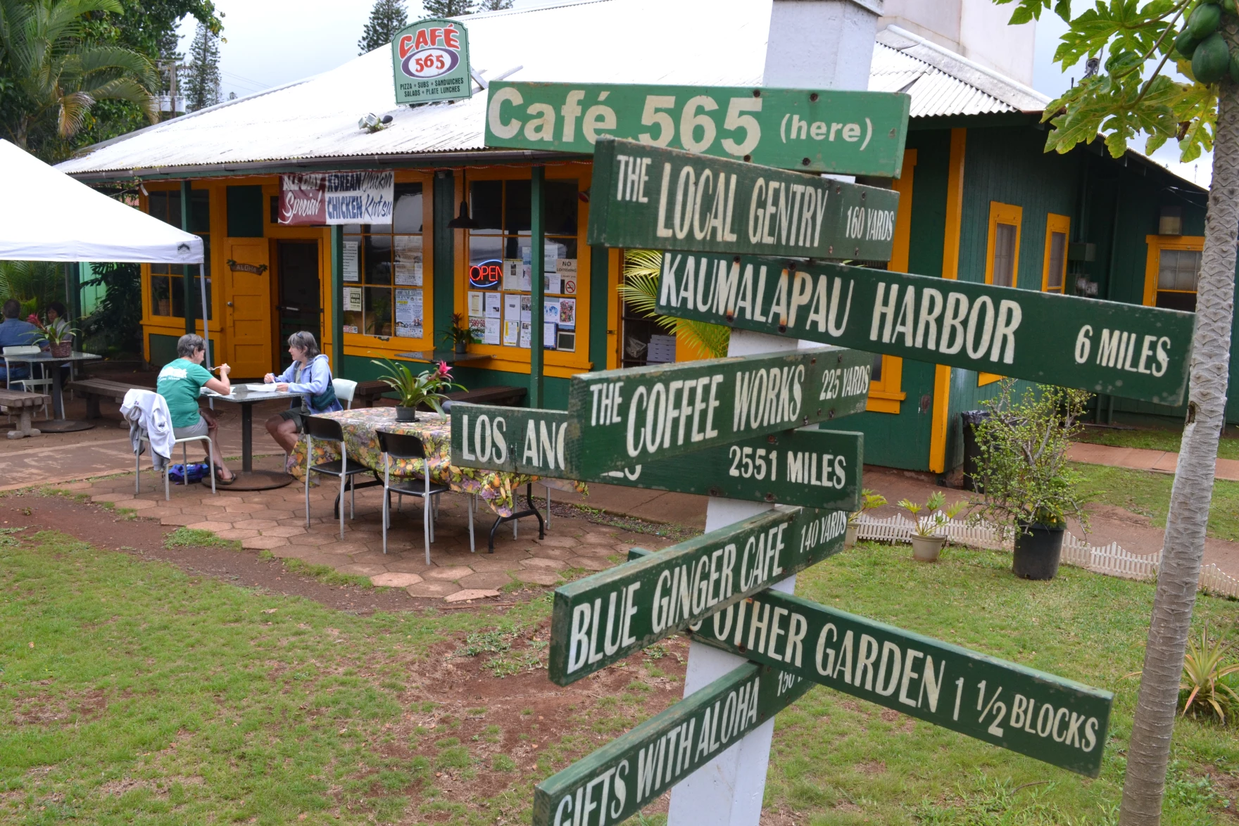 A cute green road sign telling the distance to certain destinations in front of a cafe in Lanai City.