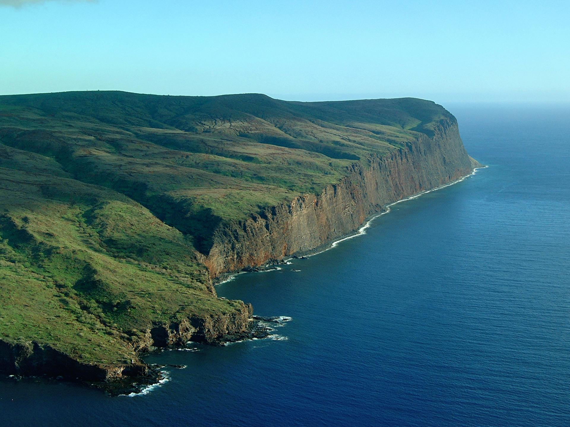 Green rugged pasture that ends in Seaside cliffs with calm blue seas below.