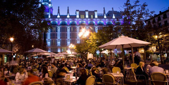 Nighttime with people sitting down eating and drinking food with tables that have umbrellas over them in the Chueca District in Madrid. 