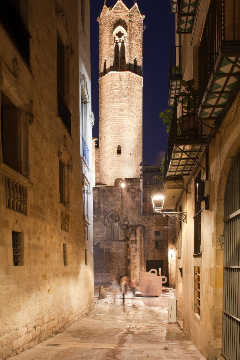 alley in the Gothic Quarter with stone streets at night with a tall medieval towers shown at the end of the alley.