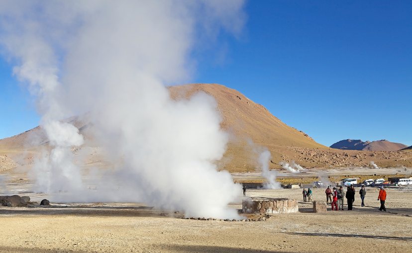 many different steam holes coming up from the ground where geysers shoot up with people watching, and a mountain in the background at El Tatio, in Northern Chile. 