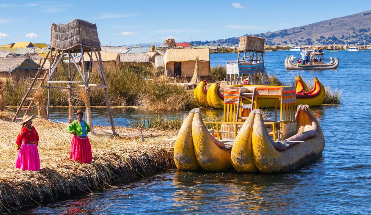 two Uru women in pink and bright colored clothes stand on a floating island made of reeds on Lake Titicaca where there is a large boat that is also made of reeds directly in front of them. 