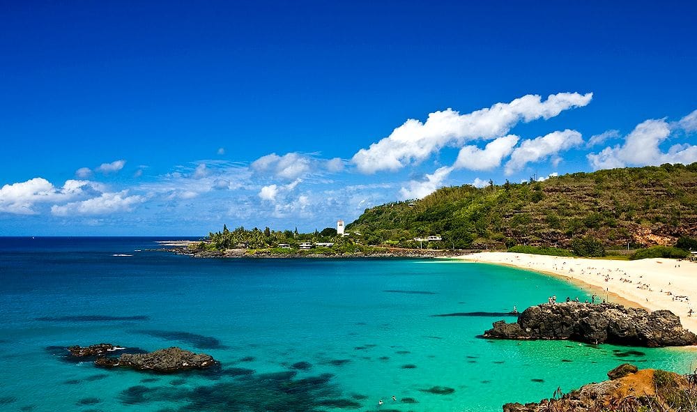 beach with clear blue waters and people standing and jumping off of a large rock in the water of Waimea Bay