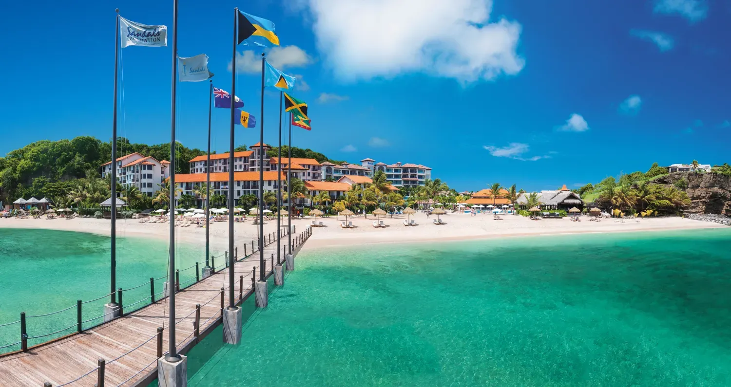 Sandals Resorts sits on the white sandy beaches of Pink Gin Beach and a pier with flags flying on it stretches out into the clear blue waters. 