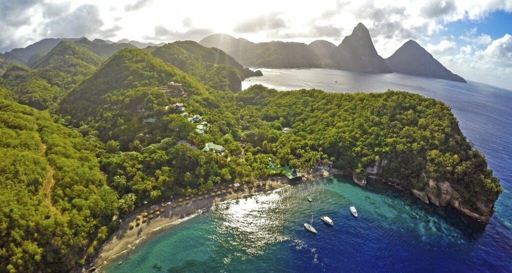 Ariel view of Anse Chastanet Beach in St. Lucia with giant mountain peaks exploding on the coastline and clear waters with white sandy beaches and sailboats anchored in front of the beach. 