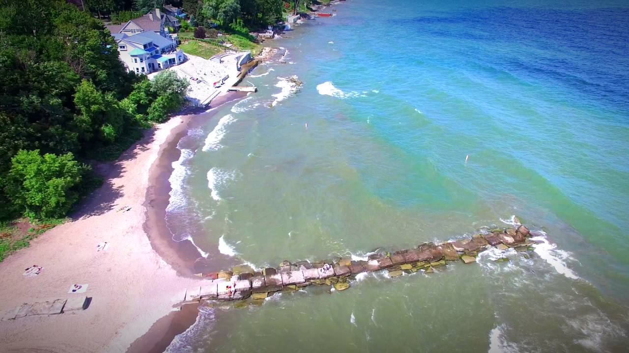 Ariel view of Bay Village Beach in Bay Village, Ohio with a rock pier with a beach on each side with clear blue waters and a house near the waters edge. 