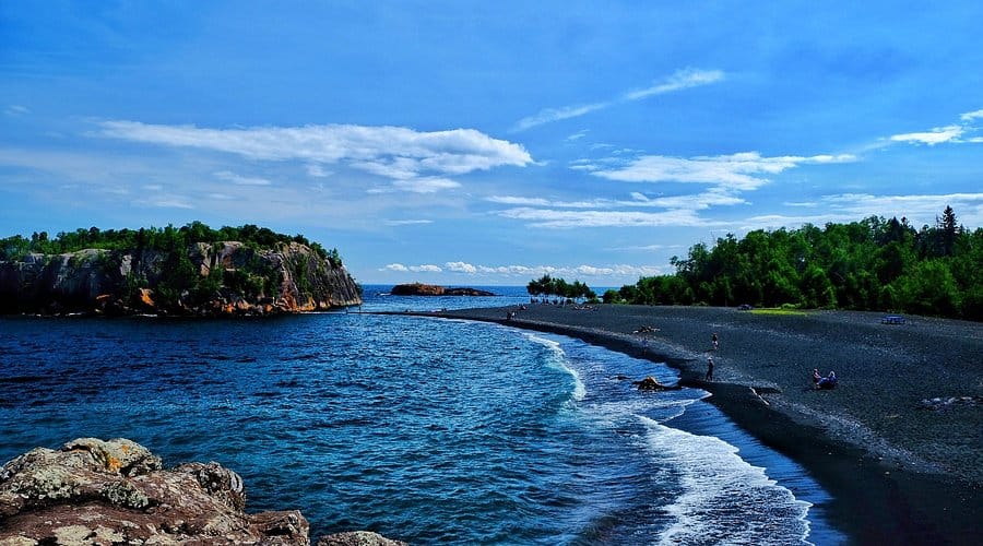 Charcoal Black sand at black beach in Silver Bay with small waves crashing the shore line and pine trees lined behind the beach. Out in the water there are jagged and rocky cliffs adding to the beauty.