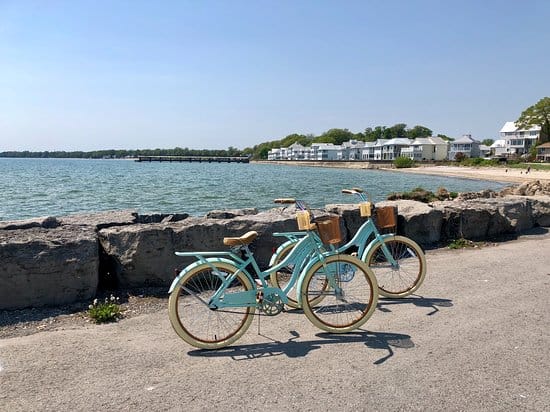 Two retro bicycles parked on the road near rocks with a view of Lake Erie in the background with a pretty beach shown near what appears to be resort condominiums.