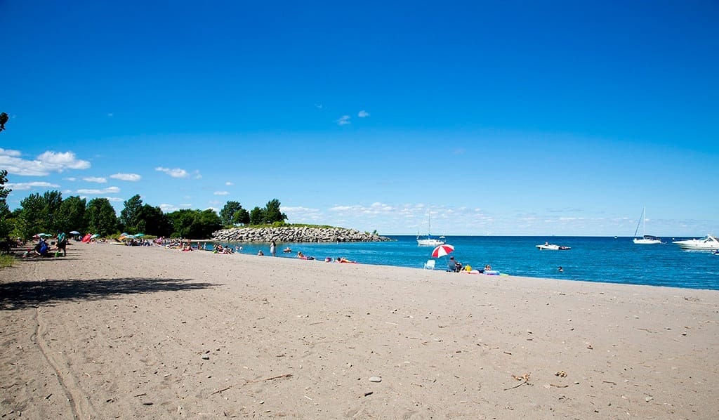 A beach with a few people enjoying a sunny day under an umbrella with boats anchored in the lake enjoying a day on the water at Fifty Point Conservation Area