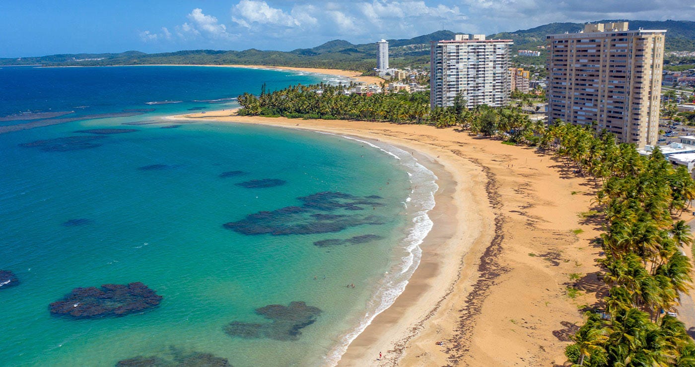 Double cover shaped beaches with tall buildings behind the beach in the morning. The beach is lined with palms behind it and the waters apear to have coral in the waters great for snorkeling. 