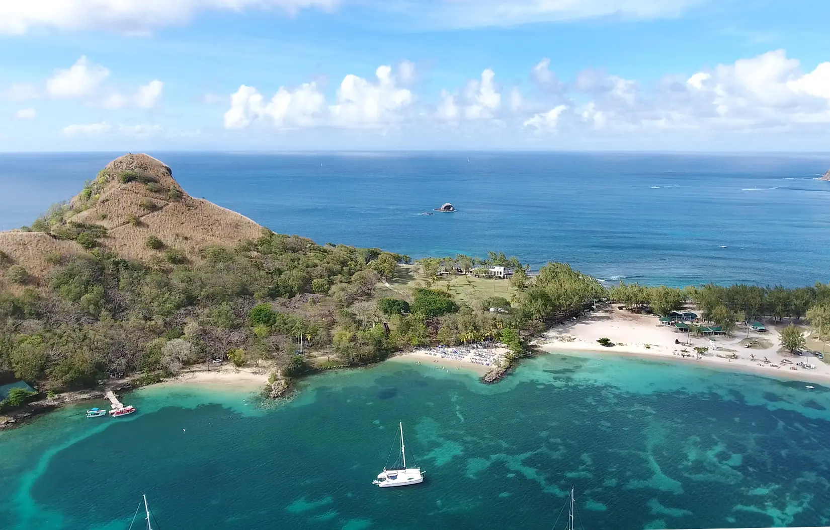 A catamaran sailboat is anchored in front of a few small beaches in clear waters in front of  mountain. The beaches have water on two sides due to being on a penensula. 