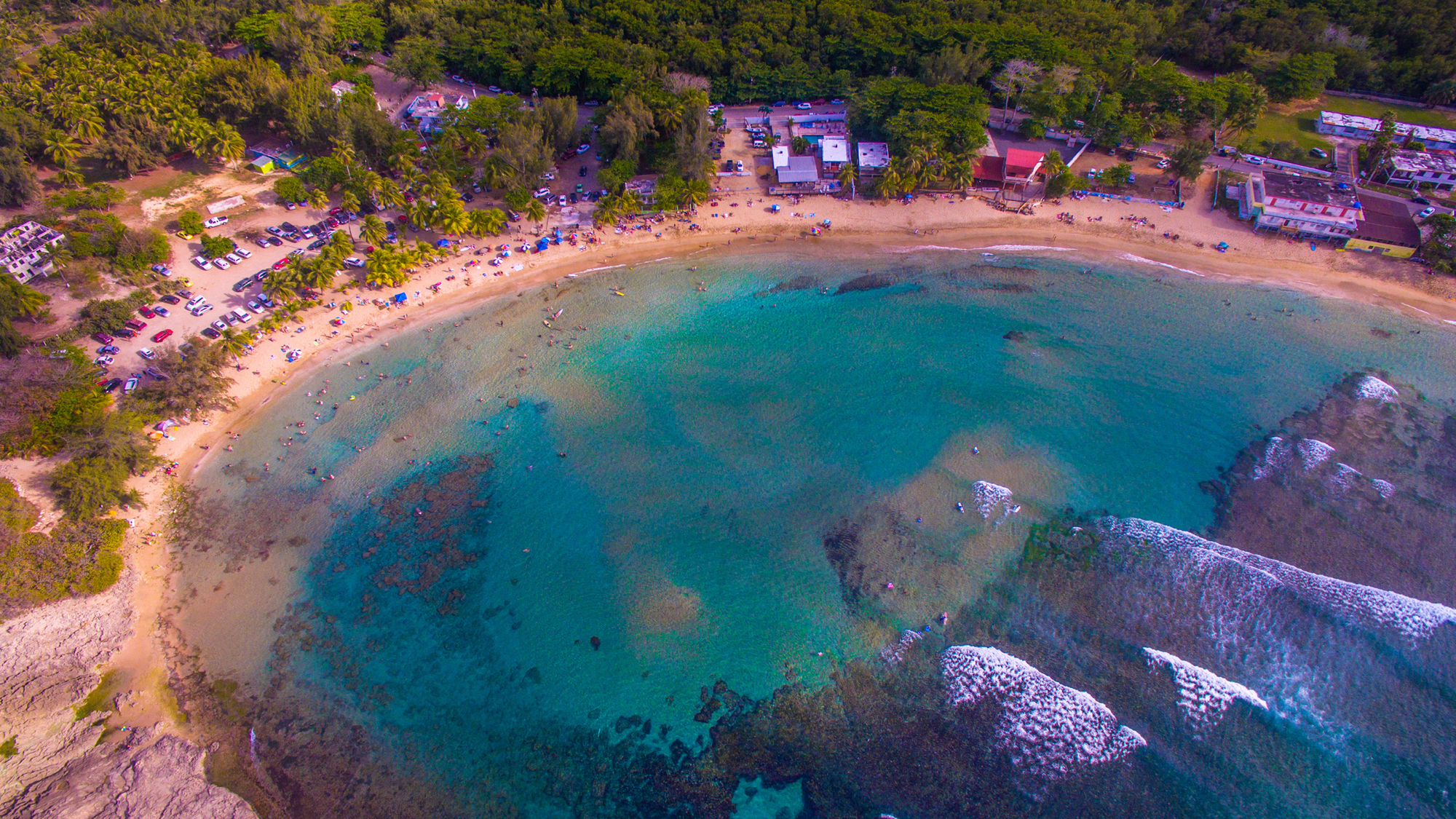 Ariel View of Playa Jobos Beach which is a horseshoe shaped cove with beach goers playing on the beach and clear waters. 