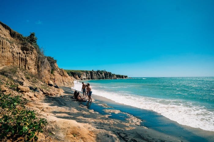 Rocky Shore lined cliffs leading to a beach below with clear blue waters and people relaxing and taking in the views. 